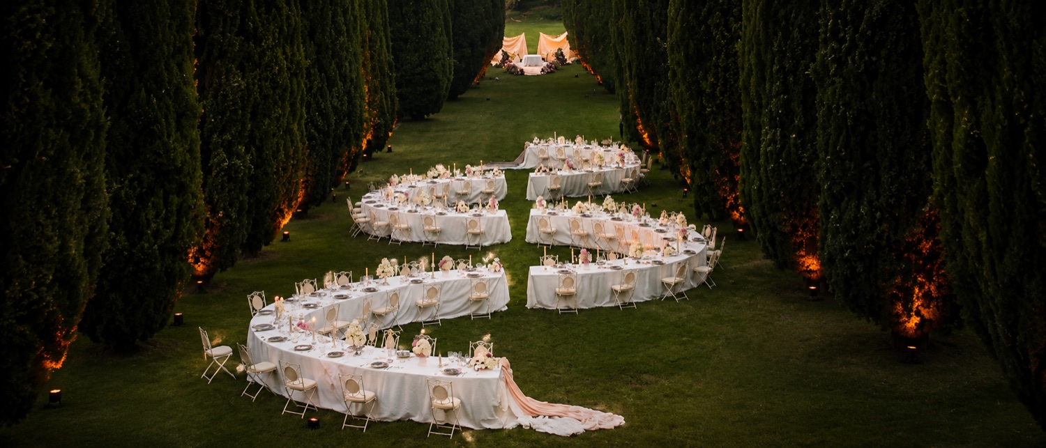 Jantar de casamento entre ciprestes ao anoitecer, mesas em curva à luz de velas